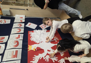 Dzieci przyklejają wycięte z papieru dłonie na szablon orła, na kształt skrzydeł.