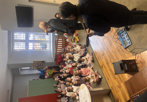 Przedszkolaki siedzą na dywanie i słuchają jak muzyk opowiada o mandolinie.