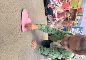 Chłopiec przekłuwa pinezką papierowe serce, na którym napisane są imiona dziewczynek..