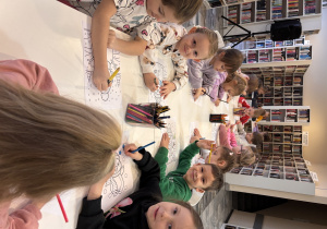 Dzieci siedzą przy długim stole i kolorują obrazki.