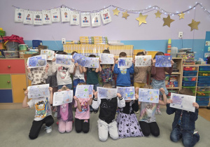 Dzieci stoją w sali przedszkolnej i zakrywają twarze zrobionymi przez siebie kartkami do Pana Premiera.