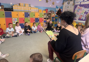 Dzieci siedzą w kole na dywanie i słuchają opowiadania czytanego przez Panią logopedę.