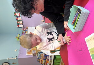 Chłopiec trzyma dłoń na kartonie, a Pani obrysowuje ją kredką.