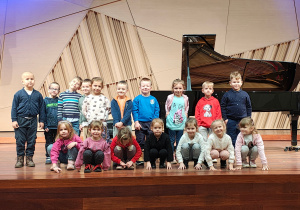 Grupa przedszkolaków pozuje do zdjęcia na scenie szkoły muzycznej. W tle widoczny fortepian.