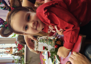 Dziewczynka w opasce renifera uśmiecha się do zdjęcia. Przed nią stoi na stoliku lampion.