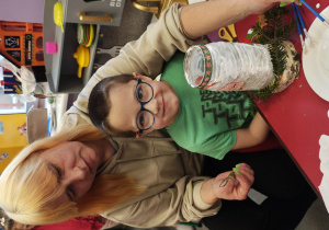 Chłopiec uśmiecha się do zdjęcia. Za nim siedzi jego mama, która pomaga mu ozdabiać lampion.