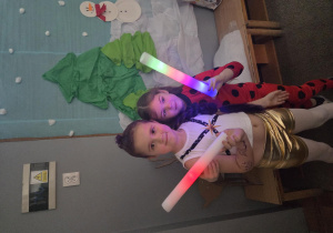 Dwie dziewczynki pozują do zdjęcia. W rękach trzymają kolorowe, świecące rurki.