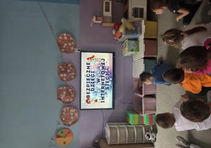 Dzieci siedząc na dywanie oglądają film edukacyjny "Bezpieczne dzieci w internetowej sieci".