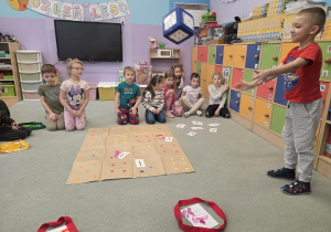 Chłopiec rzuca kolorową kostką, inne dzieci siedzą na dywanie i przyglądają się chłopcu.