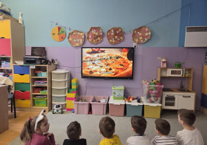 Dzieci siedzą na dywanie i oglądają film edukacyjny na temat pizzy.