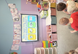 Dzieci siedzą na dywanie i rozwiązują quiz wyświetlany na ekranie multimedialnym.