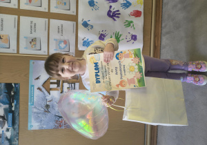 Dziewczynka pozuje do zdjęcia z dyplomem w ręku i otrzymanymi nagrodami.