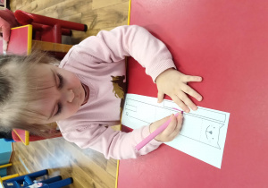 Dziewczynka siedzi przy stoliku i koloruje kota z papieru.