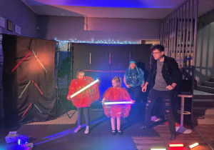 Dwie dziewczynki stoją na scenie, trzymają w rękach świecące rurki. Obok dziewczynek stoi aktor, nieco z tyłu aktorka.