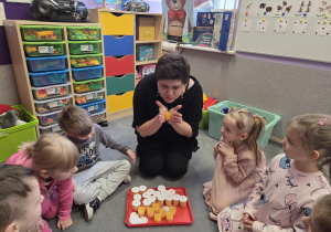 Dzieci siedzą na dywanie. Nauczycielka ogrzewa w dłoniach lody z soku pomarańczowego.