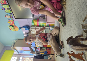 Chłopiec i dziewczynka siedzą na dywanie i bawią się figurkami dinozaurów.