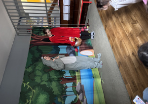 Czerwony Kapturek z wilkiem stoją na tle scenografii przedstawiającej las.