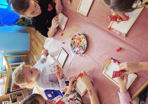 Dzieci przy stoliku ozdabiają drewniane ramki, przyklejając kolorowe guziki.