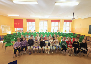 Grupa dzieci siedzi na krzesełkach w sali szkolnej, w której odbywać się będzie przedstawienie.
