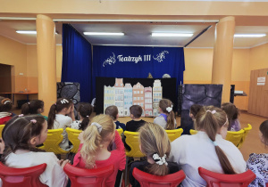 Pierwszy plan: dzieci siedzą przodem do sceny, drugi plan: scenografia na której widać kamienice.