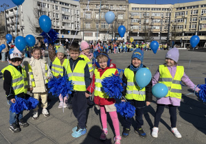 Grupa dzieci stoi na Placu Wolności. W rękach trzymają niebieskie balony i pompony.
