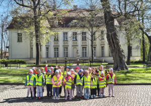 Grupa przedszkolaków w kamizelkach odblaskowych pozuje do zdjęcia na tle Pałacu Bidermana.