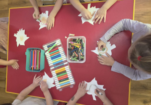 Dzieci siedzą przy czerwonym stoliku i wspólnie wykonują elementy do pracy plastycznej. Każde dziecko koloruje wycięty z papieru kształt przypominający jesienny liść, na którym znajduje się dziecięcy portret. Na środku stołu znajdują się kredki, flamastry i pastele. Dzieci samodzielnie wybierają kolory i ozdabiają swoje prace.