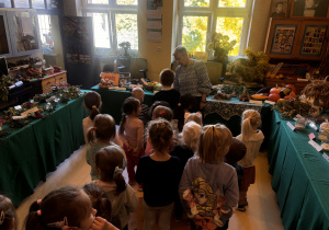 Dzieci zwiedzają wystawę „Dary jesieni” w bibliotece. Wysłuchują opowieści prowadzącej i oglądają eksponaty pochodzące z pola, lasu i sadu, prezentowane na stołach przykrytych zielonym materiałem.