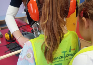 Dziecko w kamizelce odblaskowej i nausznikach ochronnych uczestniczy w pracy przy urządzeniu stolarskim. Instruktorka pomaga mu w bezpiecznym przycinaniu drewnianych elementów.