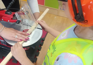 Dziecko w kamizelce odblaskowej, okularach ochronnych i nausznikach ochronnych przycina drewnianą listwę na urządzeniu stolarskim pod nadzorem instruktora.