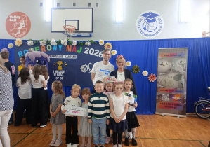 Grupa przedszkolaków pozuje do pamiątkowego zdjęcia z organizatorami kampanii „Rowerowy Maj 2026”. Dzieci trzymają czek na 10 000 zł oraz nagrody, a w tle widać dekorację z hasłem kampanii i rowerowymi motywami.