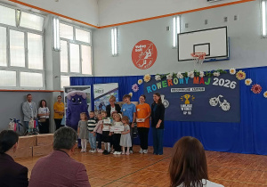 Dzieci i przedstawiciele organizatorów stoją na sali gimnastycznej przed dekoracją z napisem „Rowerowy Maj 2026 – podsumowanie kampanii”. Przedszkolaki prezentują nagrodę za zajęcie pierwszego miejsca.