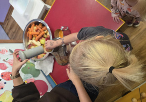 Dziecko pod opieką osoby dorosłej wkłada kawałki jabłek i marchewek do sokowirówki. Obok znajduje się metalowa miska z owocami i warzywami.